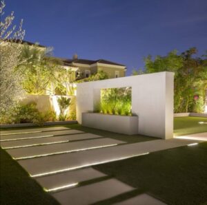Contemporary outdoor garden entrance featuring illuminated stepping stone pathway, minimalist white wall, and lush greenery creating a clean and elegant landscape design.